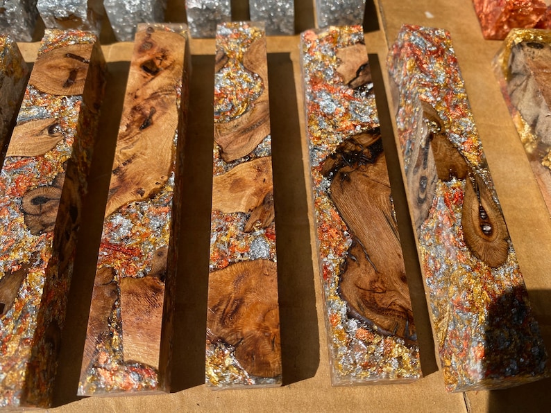 May include: Close-up of several rectangular wooden blocks with a clear resin finish. The wood has a natural grain pattern and is interspersed with silver, copper, and gold metallic flakes. The blocks are arranged side by side.