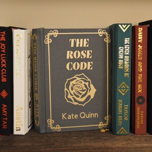 Op de afbeelding: Een boekenkast met een verscheidenheid aan boeken, waaronder "The Rose Code" van Kate Quinn, "The Seven Husbands of Evelyn Hugo" van Taylor Jenkins Reid en "Daisy Jones and the Six" van Taylor Jenkins Reid.