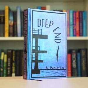 May include: A hardcover book titled "DEEP END" by Ali Hazelwood. The cover features a blue and silver design with a diving board illustration. The book is displayed in front of a bookshelf filled with various books.