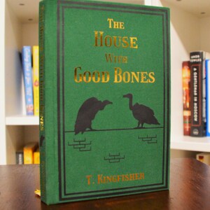 Puede incluir: Un libro de tapa dura verde titulado "The House With Good Bones" de T. Kingfisher. El libro tiene letras doradas e ilustraciones de aves. El libro est&aacute; sobre una superficie de madera.