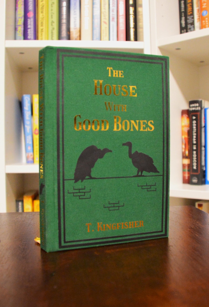 Puede incluir: Un libro de tapa dura verde titulado "The House With Good Bones" de T. Kingfisher. El libro tiene letras doradas e ilustraciones de aves. El libro est&aacute; sobre una superficie de madera.
