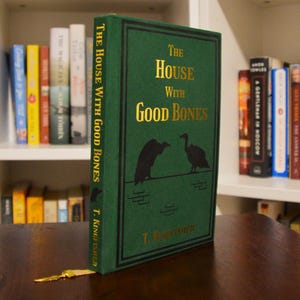 Puede incluir: Un libro de tapa dura verde titulado "The House With Good Bones" de T. Kingfisher. El t&iacute;tulo y los bordes son dorados. Dos siluetas de p&aacute;jaros negros adornan la portada. Un marcap&aacute;ginas dorado descansa sobre una superficie de madera.