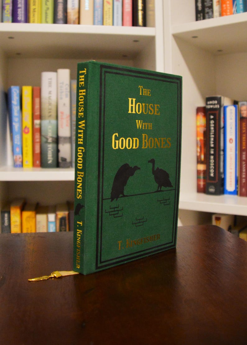 Puede incluir: Un libro de tapa dura verde titulado "The House With Good Bones" de T. Kingfisher. El t&iacute;tulo y los bordes son dorados. Dos siluetas de p&aacute;jaros negros adornan la portada. Un marcap&aacute;ginas dorado descansa sobre una superficie de madera.