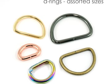May include: Assortment of D-rings in various sizes and finishes, including gold, silver, rose gold, and rainbow.