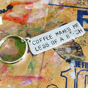 May include: A silver-colored keychain with a round ring and a rectangular tag. The tag has the phrase "COFFEE MAKES ME LESS OF A B*CH" stamped in black letters. The background is a colorful, textured surface.