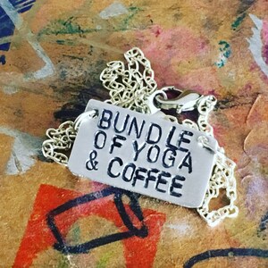 May include: Silver-toned necklace with a rectangular pendant. The pendant is stamped with the words "BUNDLE OF YOGA & COFFEE" in a bold, sans-serif font. The chain is delicate and the clasp is visible. The background is a colorful, textured surface.