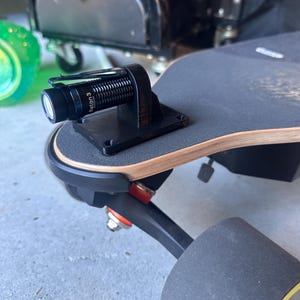 May include: A black electric skateboard with a black flashlight mounted on the deck. The flashlight is labeled "Baton 3". The skateboard has black wheels with yellow accents and a wooden deck.