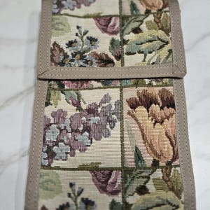 May include: A floral tapestry wallet with a beige border. The wallet features a repeating pattern of flowers in shades of purple, pink, green, and beige. The wallet is closed and appears to be made of a woven fabric.