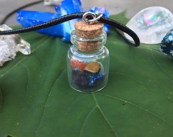May include: A small glass bottle necklace with a cork stopper. The bottle contains a variety of colorful stones, including red, orange, brown, black, blue, and green.