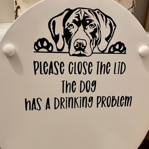 May include: White toilet seat lid with a black and white graphic of a dog's head peeking over the top. The text reads "Please close the lid, the dog has a drinking problem."