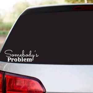 May include: A white vinyl sticker on the back window of a silver car. The sticker says "Somebody's Problem" in a cursive font.