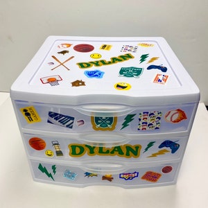 May include: White plastic three-drawer storage organizer with colorful stickers. The top drawer has a green and white name label that says "DYLAN". The middle drawer has a green and white name label that says "DYLAN". The bottom drawer has a sticker that says "SLAM DUNK".