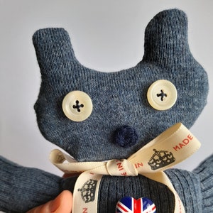 May include: A blue knitted toy with a heart-shaped patch featuring the Union Jack flag. The toy has two white buttons for eyes and a white ribbon with the text "MADE IN BRITAIN" printed on it.