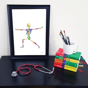 May include: A framed print of a colourful skeleton in a yoga pose. The print is on a black table with a stethoscope, a mug, and a stack of books.