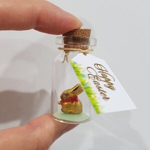 May include: A small glass bottle with a cork stopper contains a gold bunny figurine. The bottle has a green base and a tag attached that reads "Happy Easter".