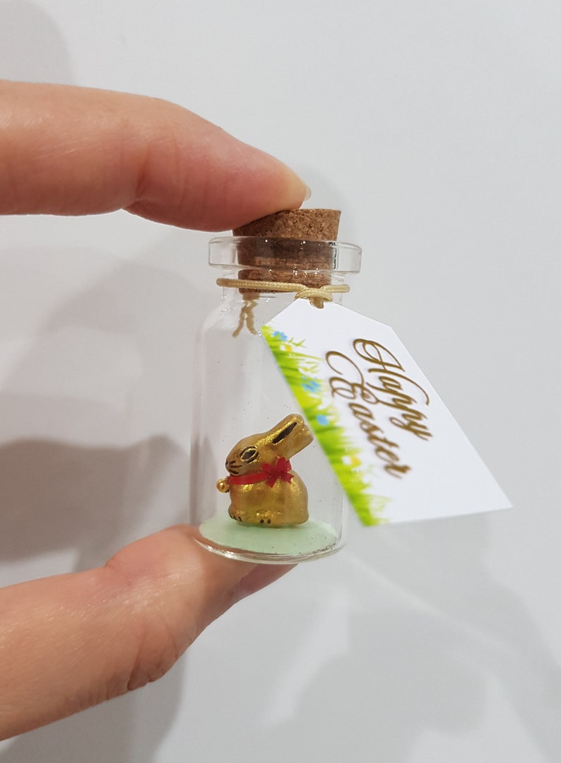 May include: A small glass bottle with a cork stopper contains a gold bunny figurine. The bottle has a green base and a tag attached that reads "Happy Easter".