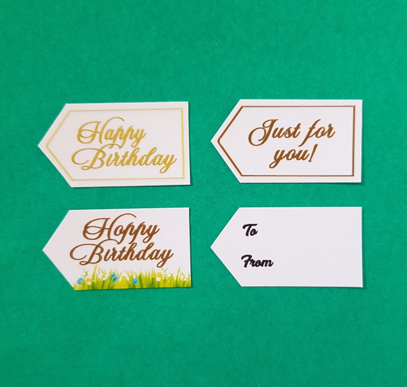 May include: Four white gift tags with gold and brown lettering. The tags have different messages: "Happy Birthday", "Just for you!" , "Happy Birthday" with a small green grass and blue flower design, and "To From".