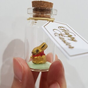 May include: A small glass bottle with a cork stopper contains a gold bunny figurine with a red bow. The bottle has a tag attached that says "Happy Easter".