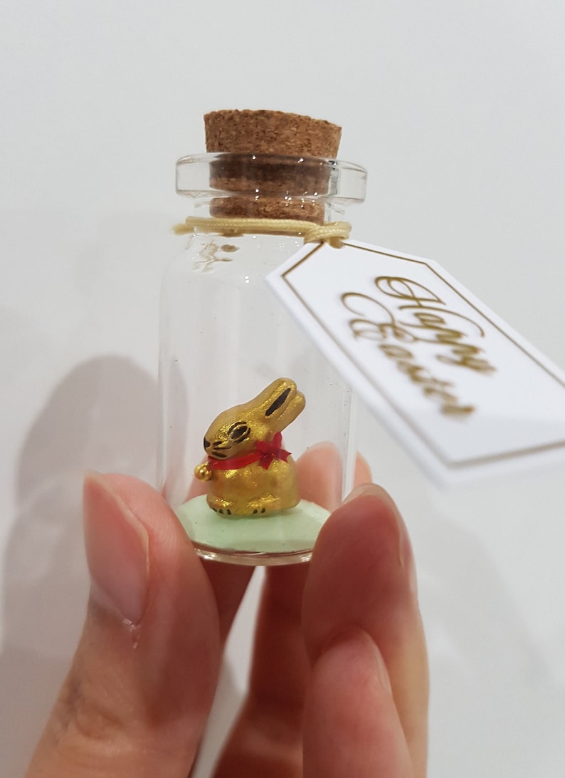 May include: A small glass bottle with a cork stopper contains a gold bunny figurine with a red bow. The bottle has a tag attached that says "Happy Easter".