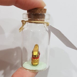 May include: A small glass bottle with a cork stopper contains a golden rabbit figurine with a red ribbon around its neck. The bottle is sitting on a green surface.