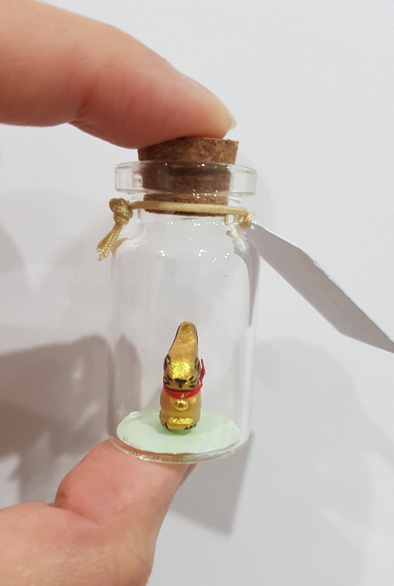 May include: A small glass bottle with a cork stopper contains a golden rabbit figurine with a red ribbon around its neck. The bottle is sitting on a green surface.