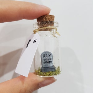 May include: A small glass bottle with a cork stopper contains a miniature tombstone with the words "RIP YOUR YOUTH" on it. The bottle is filled with green moss and a tag attached to the bottle reads "To you".