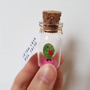 May include: A small glass jar with a cork lid contains a green olive figurine holding a red heart. The jar has a tag attached that reads "OLIVE YOU VERY MUCH!"