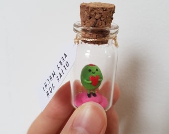 May include: A small glass jar with a cork lid contains a green olive figurine holding a red heart. The jar has a tag attached that reads "OLIVE YOU VERY MUCH!"