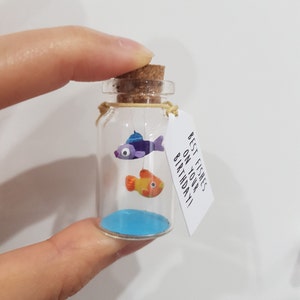 May include: A small glass jar with a cork top contains two small clay fish, one purple and one orange. The jar has a blue painted bottom and a white tag attached to the cork that reads "Best fishes on your birthday!"