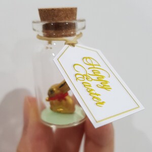 May include: A small glass bottle with a cork stopper and a gold bunny figurine inside. The bottle has a white tag tied around it that says "Happy Easter" in gold lettering.