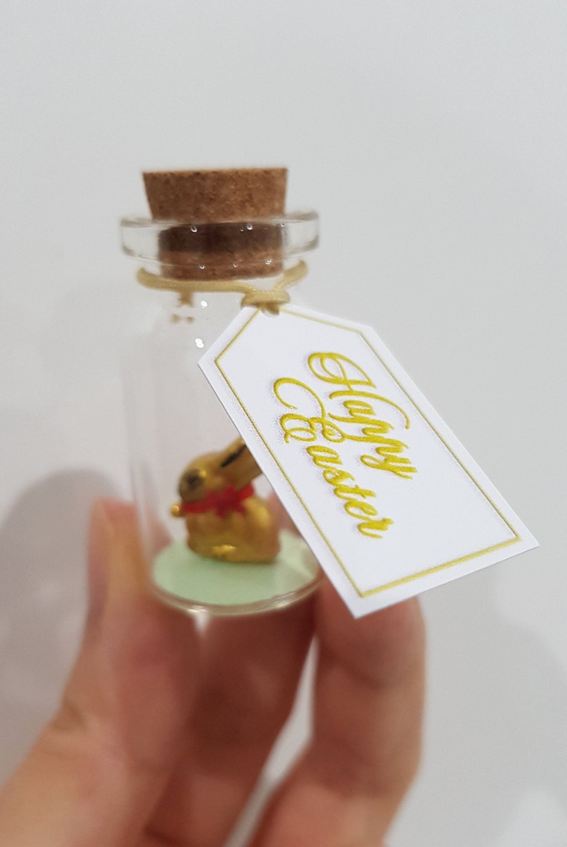 May include: A small glass bottle with a cork stopper and a gold bunny figurine inside. The bottle has a white tag tied around it that says "Happy Easter" in gold lettering.