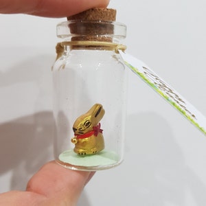 May include: A small glass bottle with a cork stopper contains a gold rabbit figurine wearing a red bow. The bottle has a green base and a tag that says "Good Luck".