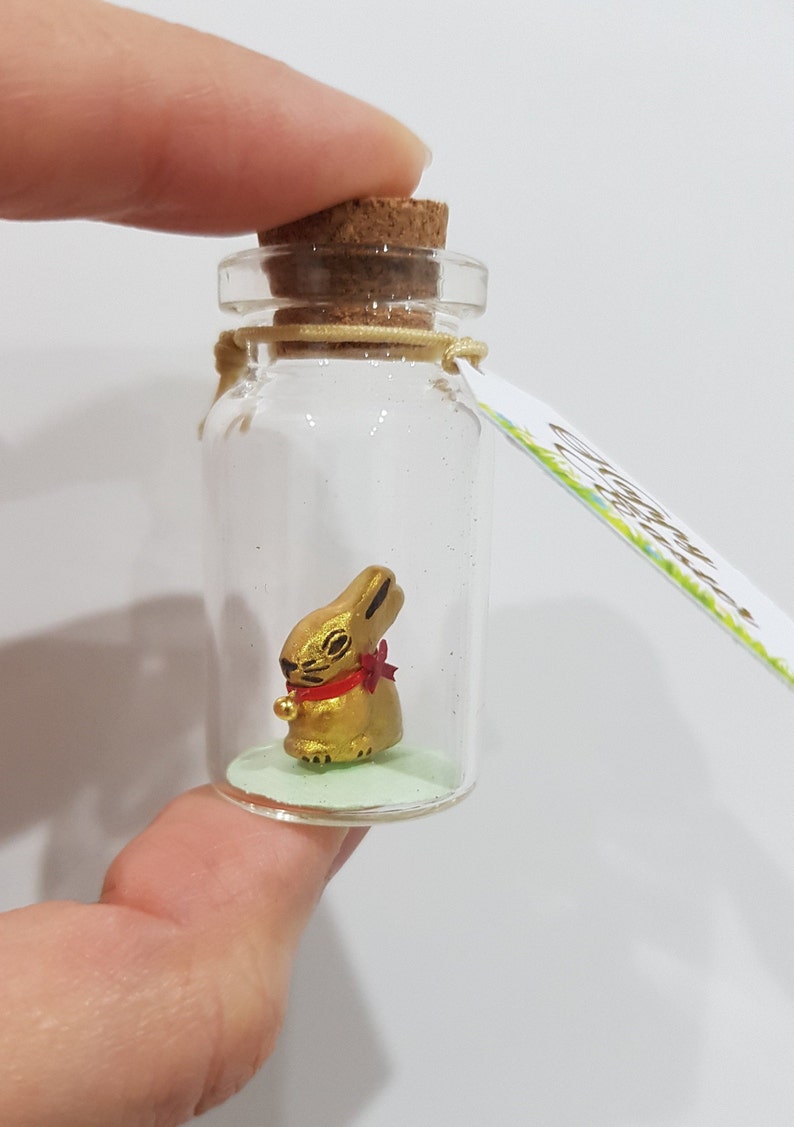 May include: A small glass bottle with a cork stopper contains a gold rabbit figurine wearing a red bow. The bottle has a green base and a tag that says "Good Luck".