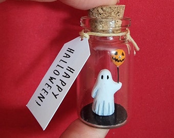 May include: A small glass bottle with a cork stopper contains a white ghost figurine holding a small orange jack-o'-lantern balloon. A white tag attached to the bottle reads "Happy Halloween!"