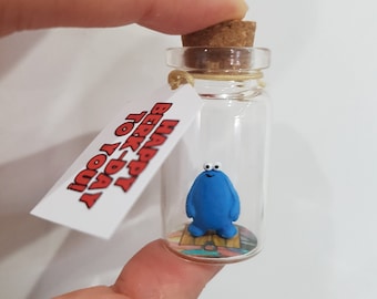 May include: A small glass bottle with a cork stopper contains a blue figurine with white eyes. A white tag attached to the bottle reads "Happy Birthday to You!"