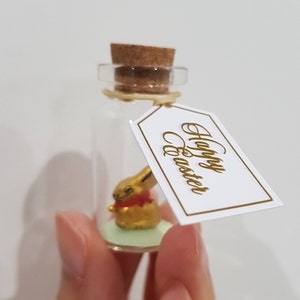 May include: A small glass bottle with a cork stopper contains a gold bunny figurine. A white tag with gold lettering reads "Happy Easter".