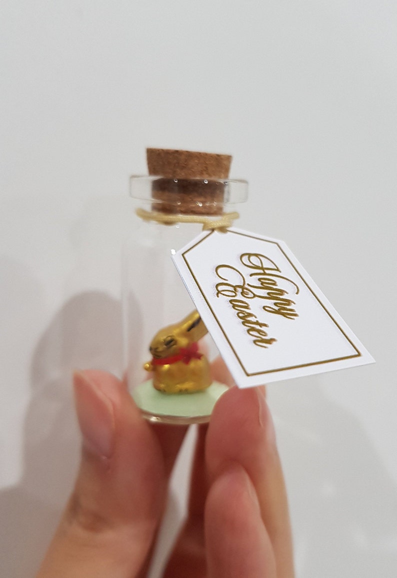 May include: A small glass bottle with a cork stopper contains a gold bunny figurine. A white tag with gold lettering reads "Happy Easter".