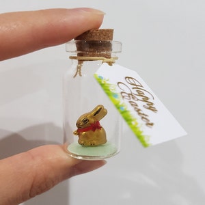 May include: A small glass bottle with a cork stopper contains a gold rabbit figurine with a red bow. A tag attached to the bottle reads "Happy Easter".