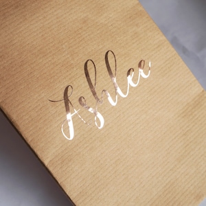 May include: A brown paper gift bag with the name "Ashlee" written in gold foil lettering.