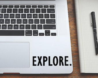 May include: A silver laptop computer with a black keyboard and a black trackpad. The laptop is open and the screen is black. The laptop is sitting on a wooden table with a black pen and a notebook. The laptop has a sticker on the bottom right corner that says "EXPLORE."