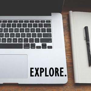 May include: A silver laptop computer with a black keyboard and a black trackpad. The laptop is open and the screen is black. The laptop is sitting on a wooden table with a black pen and a notebook. The laptop has a sticker on the bottom right corner that says "EXPLORE."