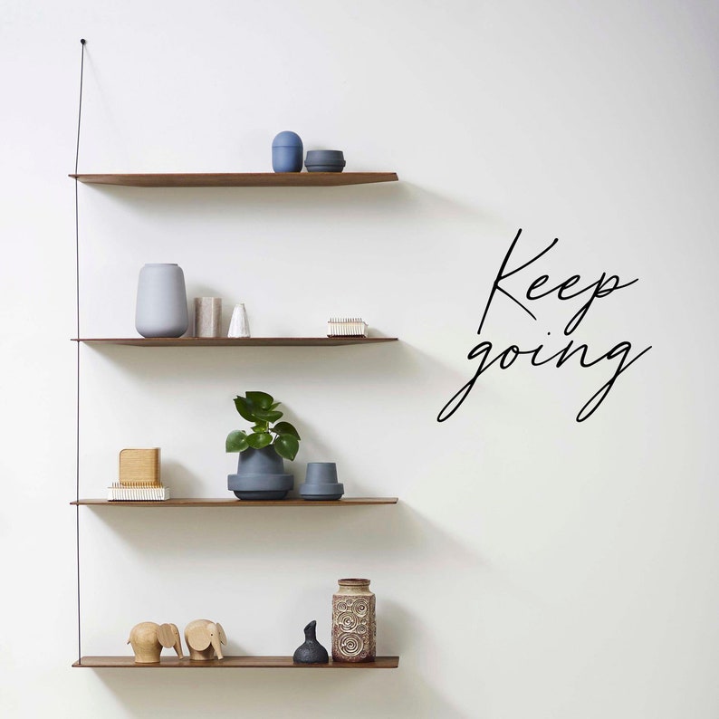 K&ouml;nnte beinhalten: Drei Holzregale mit verschiedenen Dekorationsartikeln, darunter eine Pflanze, Vasen und Figuren. Die Regale sind an einer wei&szlig;en Wand montiert, auf der der Text "Keep going" in schwarzer Kursivschrift steht.