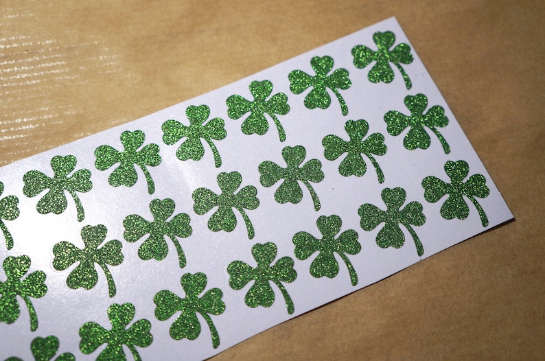 May include: A sheet of white paper with green glitter shamrock stickers. The shamrocks are arranged in rows and are all the same size.