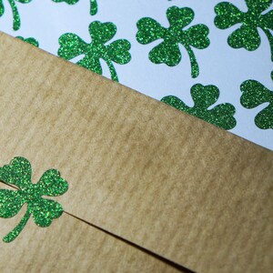 May include: A brown paper envelope with a green glitter shamrock on the front. The envelope is lying on top of a sheet of paper with green glitter shamrocks.