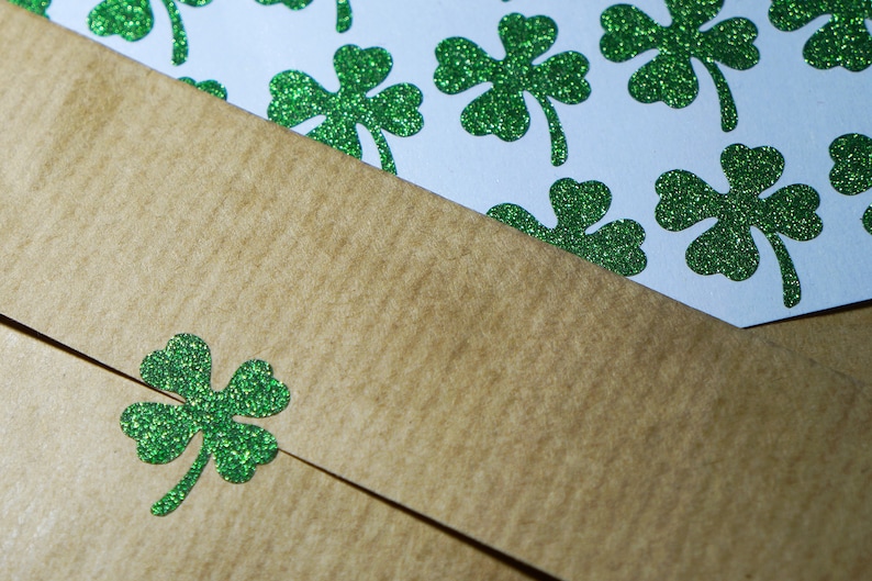 May include: A brown paper envelope with a green glitter shamrock on the front. The envelope is lying on top of a sheet of paper with green glitter shamrocks.