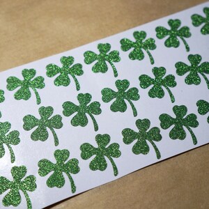 May include: A sheet of white paper with 20 green glitter shamrock stickers.