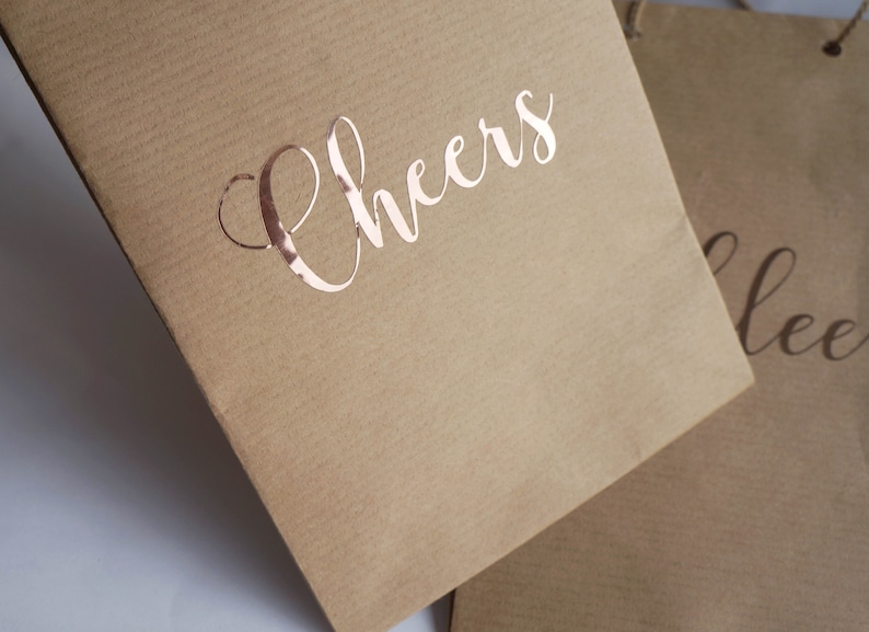 May include: Two brown paper gift bags with handles. The bag on the left has the word "Cheers" printed in rose gold foil. The bag on the right has the word "Cheers" printed in black.