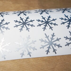 May include: A sheet of white paper with twelve silver snowflake stickers. The snowflakes are all the same design, with six points and a slightly textured surface.