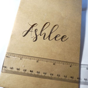 May include: A brown paper gift bag with a gold string handle. The name "Ashlee" is printed in cursive on the front of the bag.