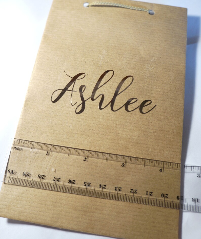 May include: A brown paper gift bag with a gold string handle. The name "Ashlee" is printed in cursive on the front of the bag.